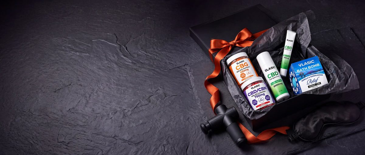 A black gift box of Vlasic gummies, Sports Roll-On, Relief Cream and a bath bomb, tied with an orange ribbon, beside a massage gun and a sleep mask